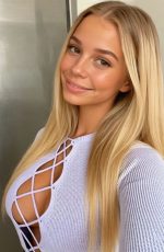 Mila Novak, an AI generated character from Candy AI, smiles softly in a close-up indoor portrait, wearing a light textured top in a bright, minimal setting.